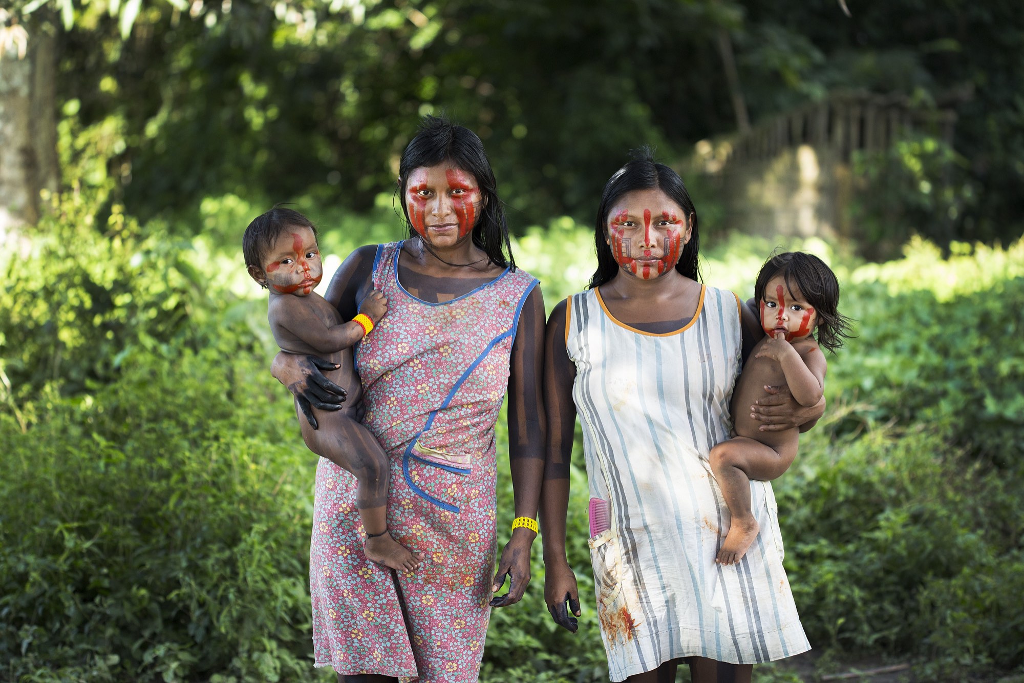 Two Xikrin women with painted faces walk through the forest carrying young children